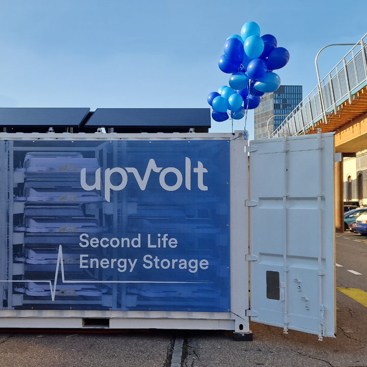 upVolt Second-Life-Energiespeicher im Container: geöffnete Front mit gestapelten Batteriemodulen, Solarmodule auf dem Dach, blaue Ballons zur Einweihung.