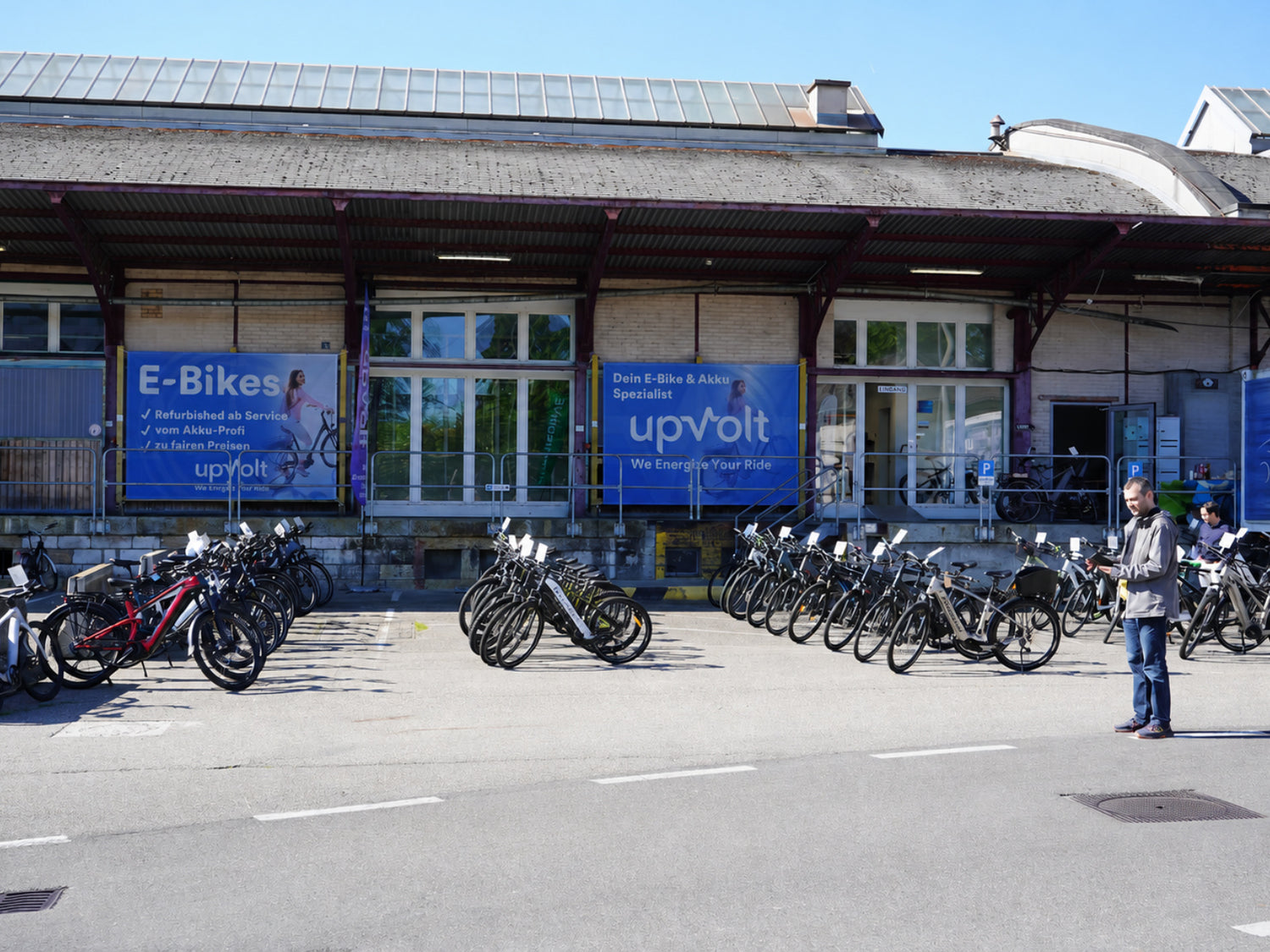 Aussenansicht der upVolt-Werkstatt in Basel. Alte Zoll-Halle mit blauen upVolt-Bannern und einer grossen Vielfalt sn  E-Bikes auf dem Vorplatz.