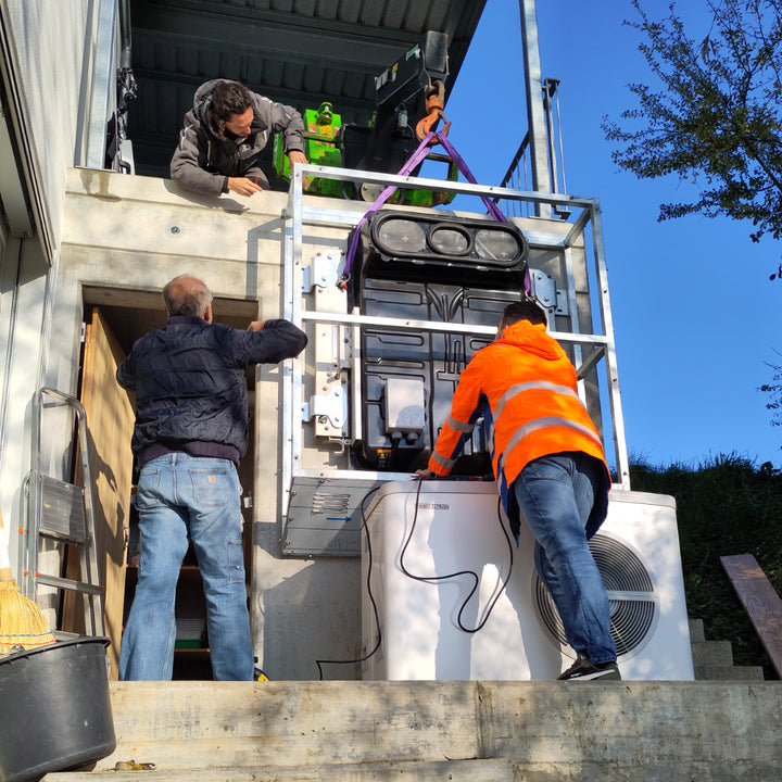 Wandmontage einer upVolt Power Unit (40 kWh): Das Team hebt das Batteriepaket per Kran in den Wandrahmen, daneben eine Wärmepumpe.