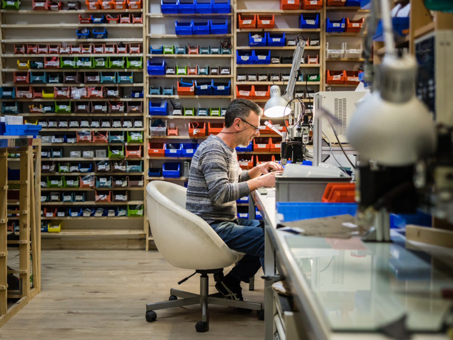 upVolt-Techniker an der Werkbank beim Reparieren, dahinter eine Regalwand mit sortierten Ersatzteil-Boxen.